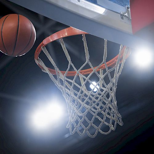 The image shows a basketball approaching a hoop, framed by bright stadium lights in the background.