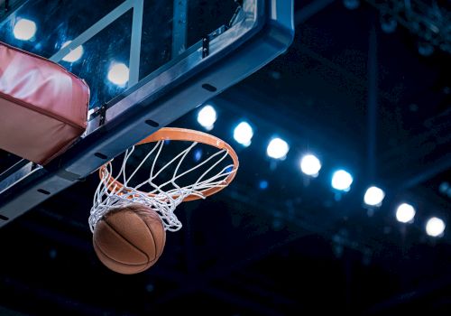 A basketball going through a hoop with a backboard, set against a backdrop of bright arena lights.