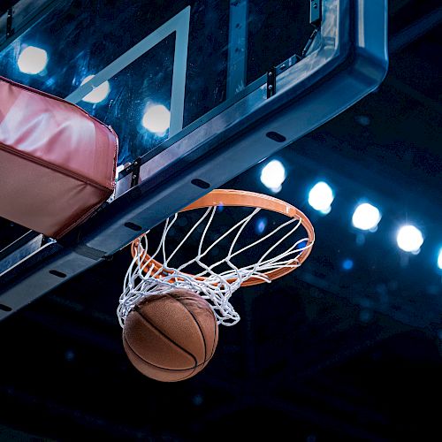 A basketball going through a hoop with a backboard, set against a backdrop of bright arena lights.