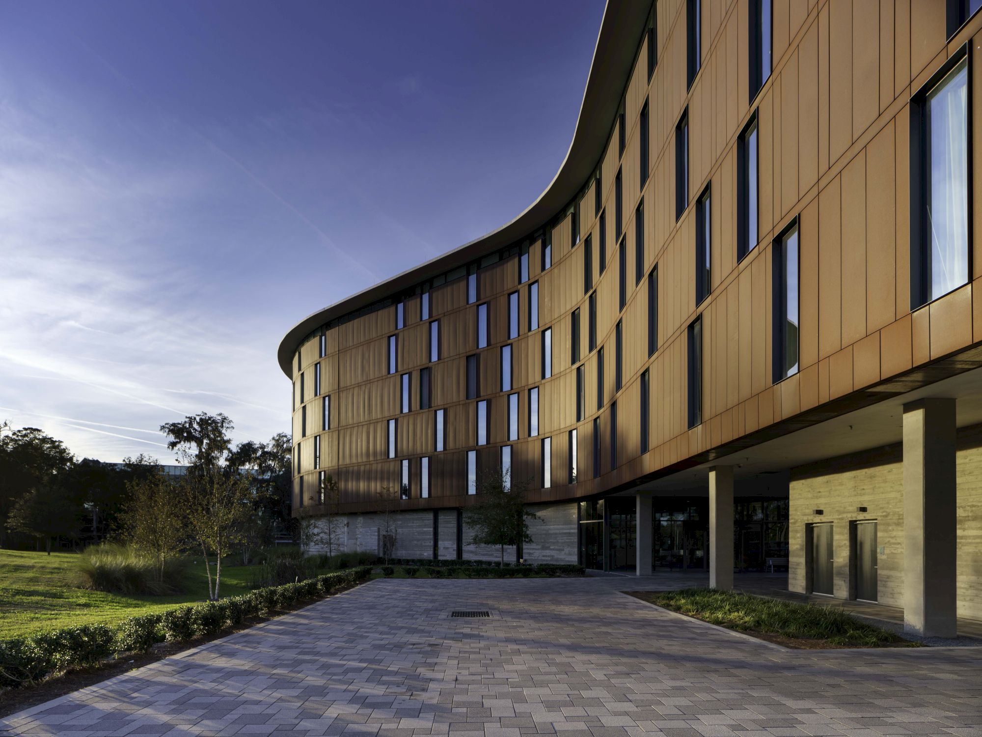 A modern, curved building with a brown facade, large windows, and an entrance area, surrounded by greenery under a clear sky.