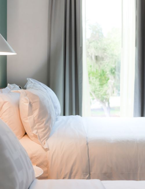 The image shows a cozy bedroom with neatly arranged white pillows and bedsheets, next to a window with curtains drawn aside.