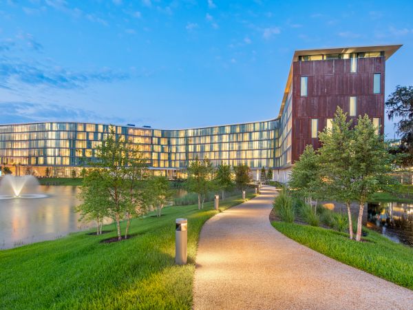A modern building by a pond with fountains, surrounded by trees and a lit pathway at dusk.