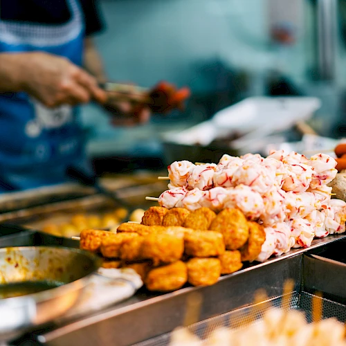 An assortment of skewered street food, including seafood, meatballs, and sausages, is displayed at a food stall.
