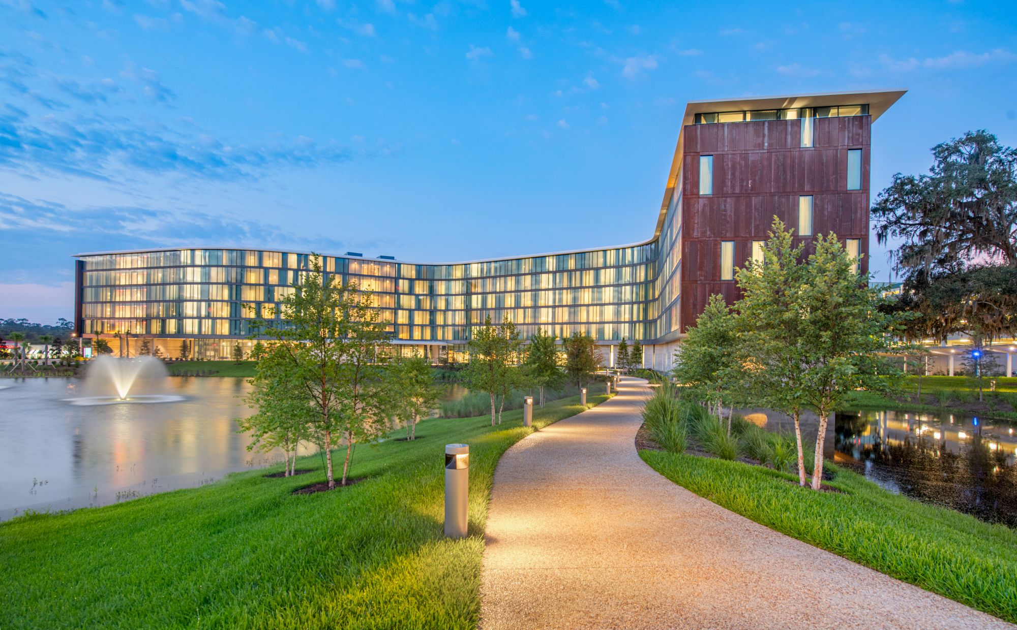 A modern building by a pond with a fountain, surrounded by trees and a winding path, under a blue sky at dusk.