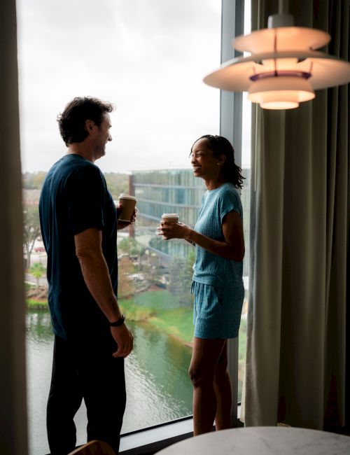 Two people are standing by a window, holding cups and talking, with an outdoor view and a ceiling light in the room.