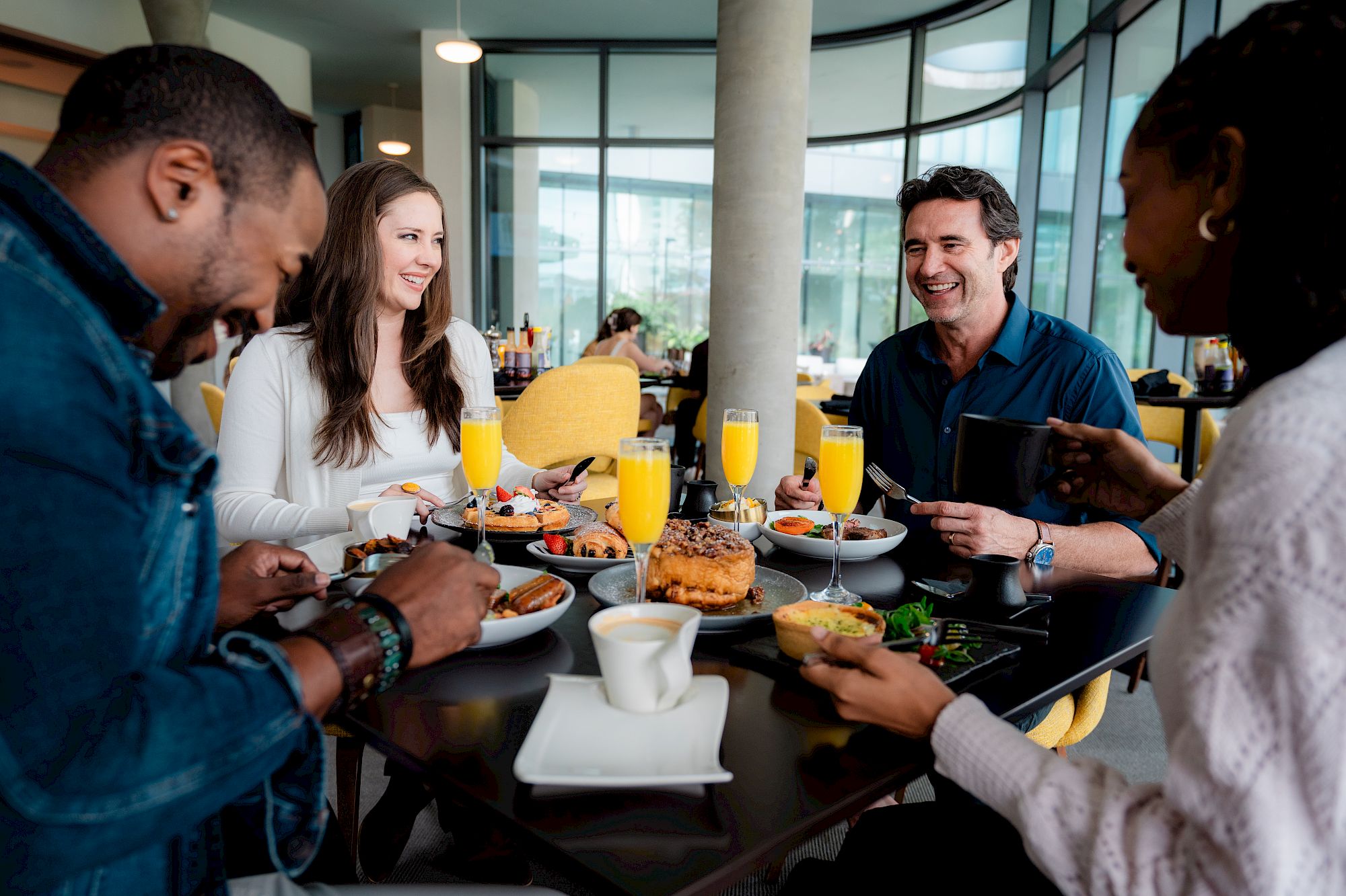 A group of people enjoying a meal with drinks at a restaurant table, smiling and conversing in a modern, bright setting.