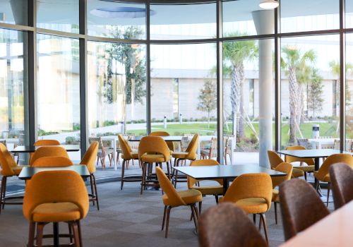 A spacious, modern seating area with yellow chairs and tables near large windows, offering a view of greenery outside.