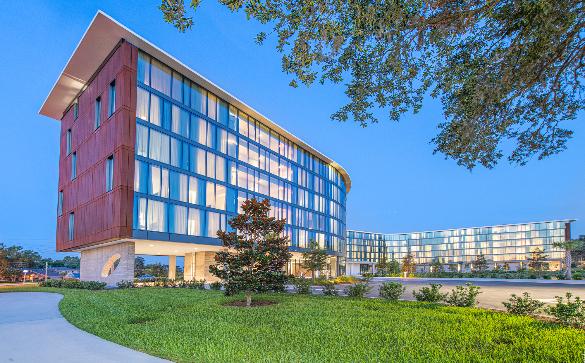 A modern building with large glass windows and a landscaped foreground under a clear, blue sky at dusk.