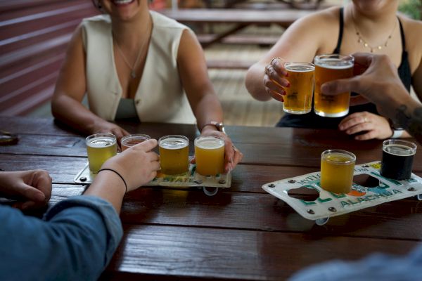 People are enjoying a variety of beers at a wooden table, with some toasting and others holding beer flights.