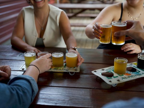 People are enjoying a variety of beers at a wooden table, with some toasting and others holding beer flights.