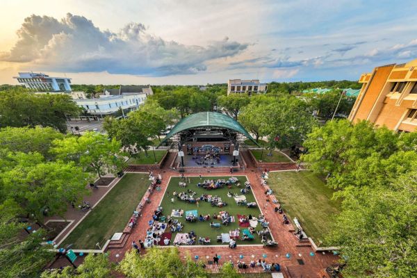 An aerial view of a park with a stage and audience seated on the grass, surrounded by trees and buildings at sunset.