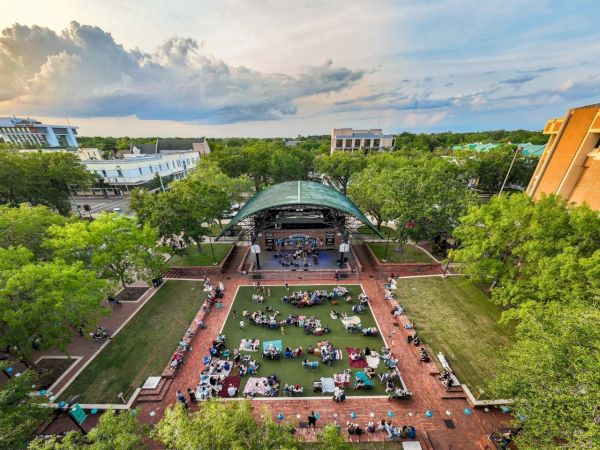 An aerial view of a park with a stage and audience seated on the grass, surrounded by trees and buildings at sunset.