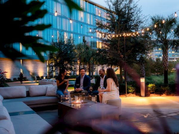 A group of people are sitting outdoors, socializing around a fire pit with string lights above, surrounded by trees and modern buildings.