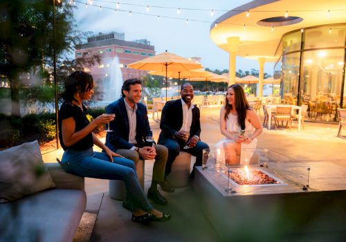 A group of people enjoy an evening outdoors near a fire pit, with string lights and a modern building in the background, creating a cozy ambiance.