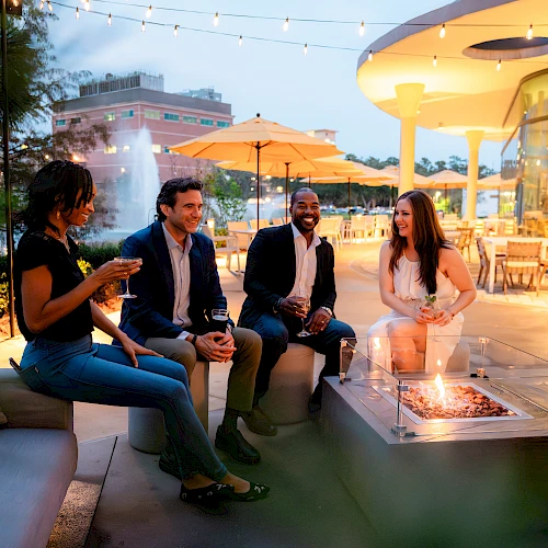 A group of people enjoy an evening outdoors near a fire pit, with string lights and a modern building in the background, creating a cozy ambiance.