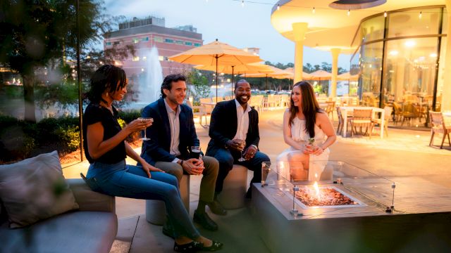 A group of people enjoy an evening outdoors near a fire pit, with string lights and a modern building in the background, creating a cozy ambiance.
