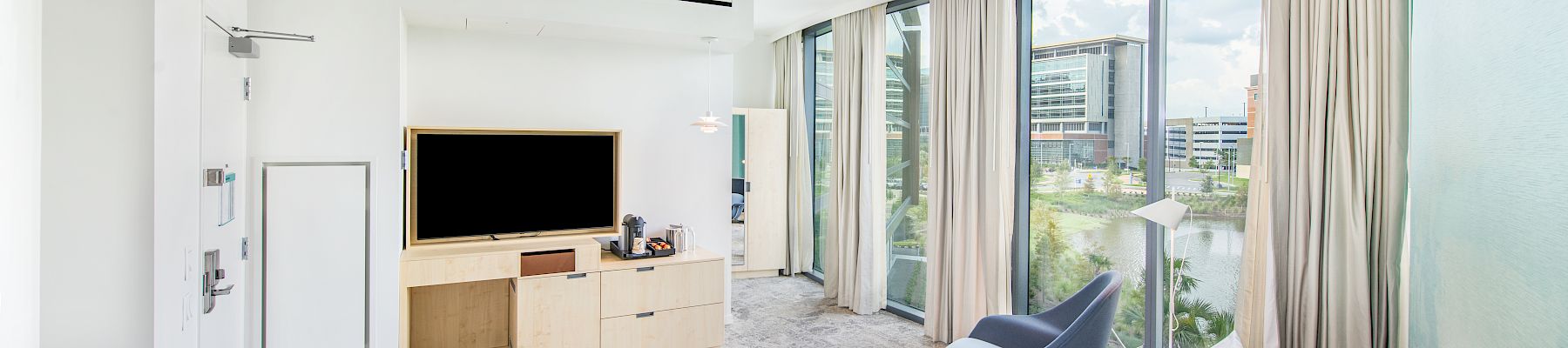 A modern hotel room with a bed, TV, chair, and large windows offering a view of nearby buildings and greenery.