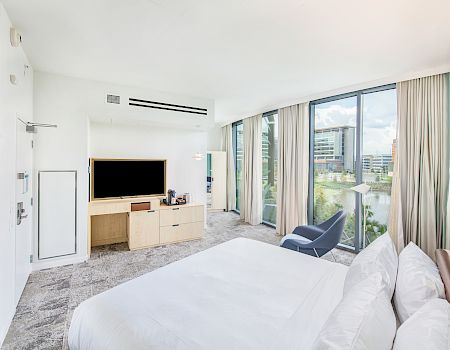 A modern hotel room with a bed, TV, chair, and large windows offering a view of nearby buildings and greenery.