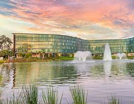 The image shows a modern glass building by a lake with fountains under a colorful sunset sky.