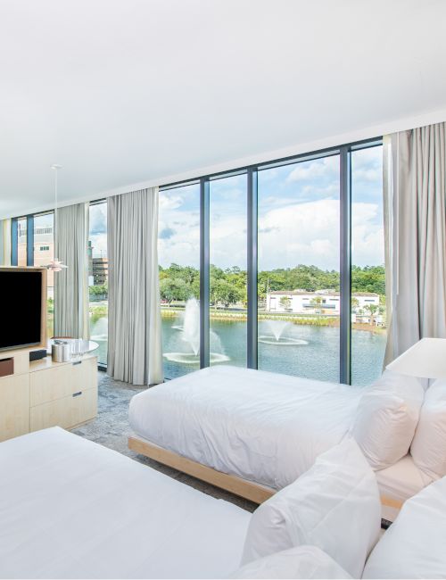 A bright hotel room with two white beds, a wooden wardrobe and TV, large floor-to-ceiling windows overlooking a lake and trees. End.