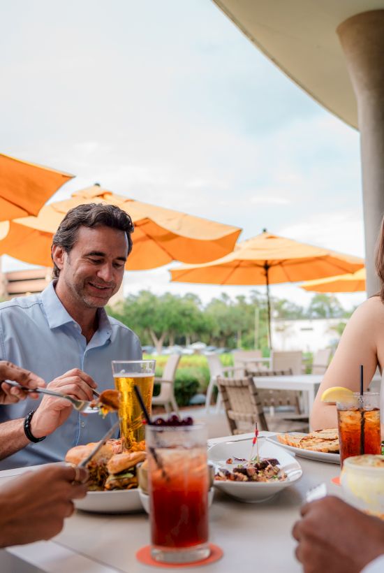 A group of four friends dining at an outdoor cafe, sharing drinks and plates under orange umbrellas, enjoying conversation and food.