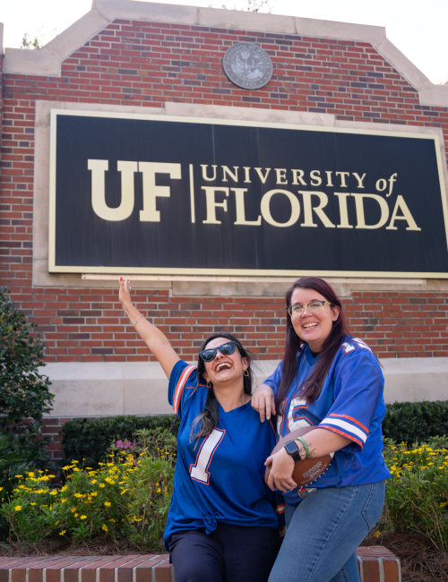 Two students pose cheerfully in blue shirts in front of a UF sign at the University of Florida, smiling and pointing up at the campus landmark.
