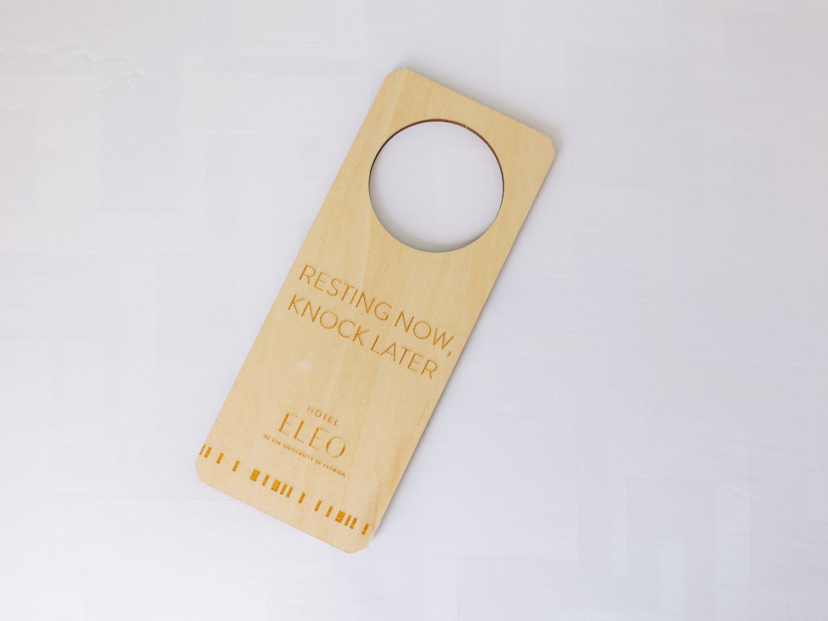 The image shows a wooden door hanger with a circular hole, reading "Resting now, knock later" and "Hotel LEO" at the bottom.