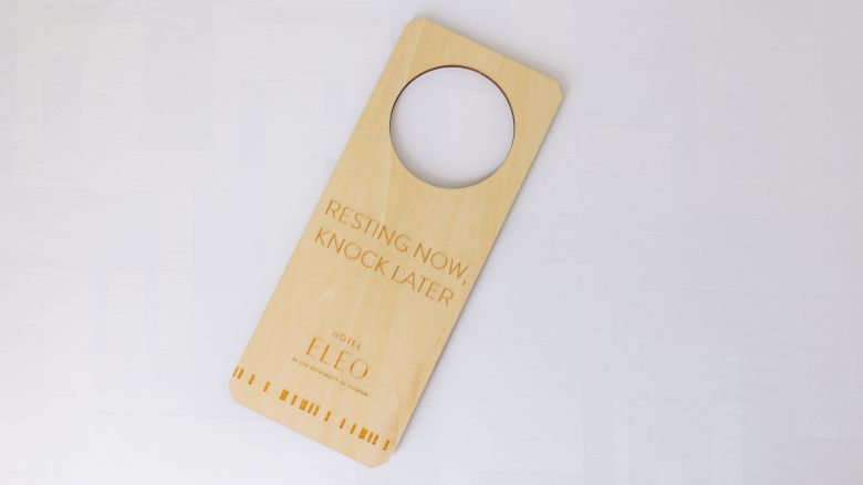 The image shows a wooden door hanger with a circular hole, reading "Resting now, knock later" and "Hotel LEO" at the bottom.