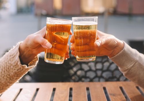 Two people clinking glasses filled with a light beverage, sitting at a wooden table in an outdoor setting.