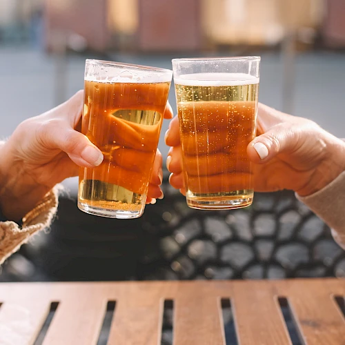 Two people clinking glasses filled with a light beverage, sitting at a wooden table in an outdoor setting.