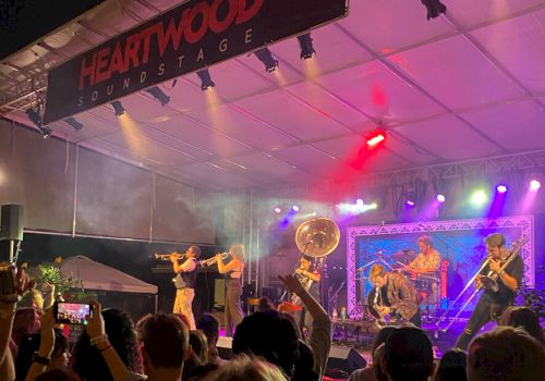 A live band performs on stage under a tent with colorful lights, as a crowd watches and raises phones to capture the moment.