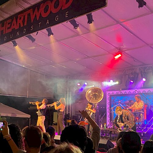 A live band performs on stage under a tent with colorful lights, as a crowd watches and raises phones to capture the moment.