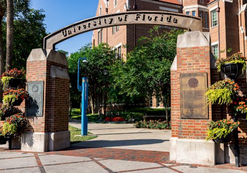 An entrance archway with "University of Florida" inscribed, surrounded by brick pillars and lush greenery in the background.
