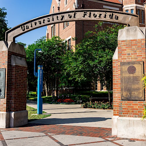 An entrance archway with "University of Florida" inscribed, surrounded by brick pillars and lush greenery in the background.