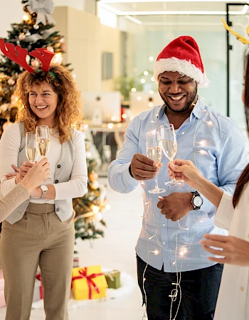 A cheerful group of coworkers in festive hats clinking glasses at an office holiday party beside a decorated Christmas tree.