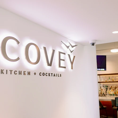 A chic restaurant interior with a white wall displaying the Covey Kitchen & Cocktails sign, bar area visible in the background.