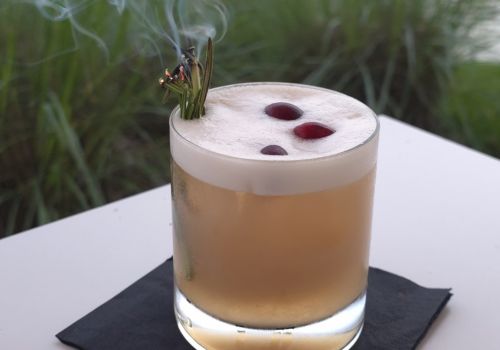 A smoky cigar rests on the rim of a glass of light brown cocktail, with a blurred fountain and buildings in the background.