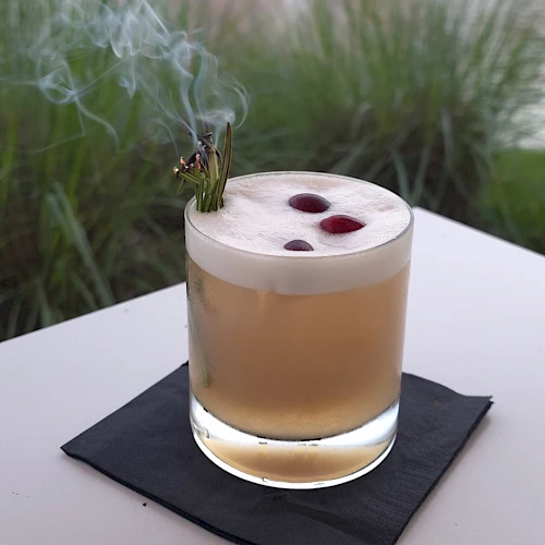 A smoky cigar rests on the rim of a glass of light brown cocktail, with a blurred fountain and buildings in the background.