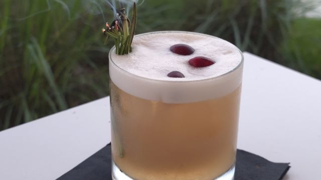 A smoky cigar rests on the rim of a glass of light brown cocktail, with a blurred fountain and buildings in the background.
