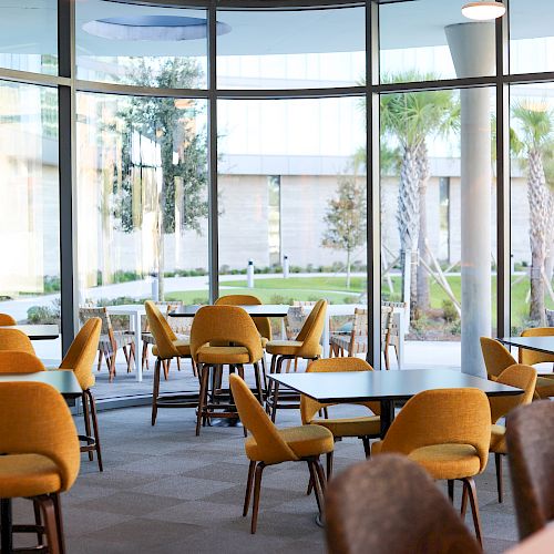 A spacious, modern seating area with yellow chairs and tables near large windows, offering a view of greenery outside.