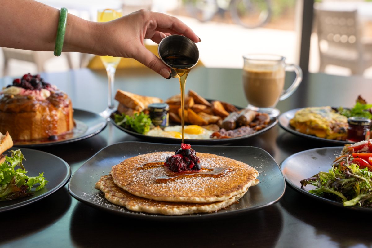 A person pours syrup over a stack of pancakes with berries, surrounded by a spread of savory plates, salads, and drinks on a table, breakfast feast.
