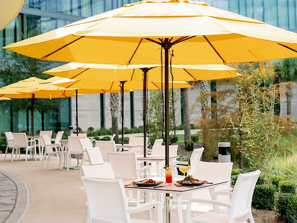 An outdoor patio seating area with white chairs and tables under bright yellow umbrellas, beside a modern glass office building.