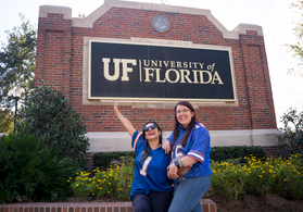 Two students pose in UF blue jerseys in front of a University of Florida sign, smiling under a sunny sky.