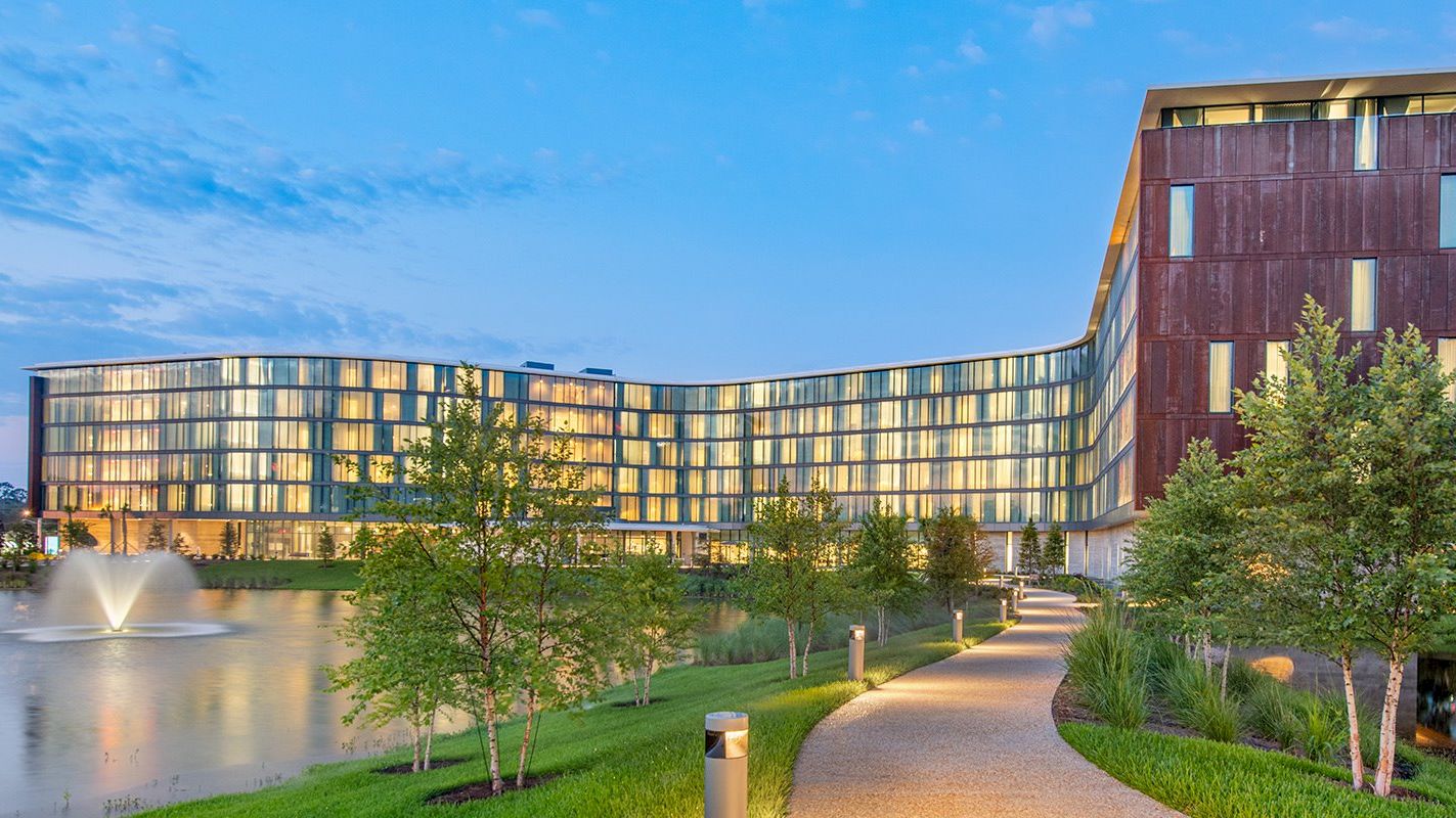 The image shows a modern, multi-story building with glass windows, a curved design, a water fountain, and a landscaped path with trees and lights ending the sentence.