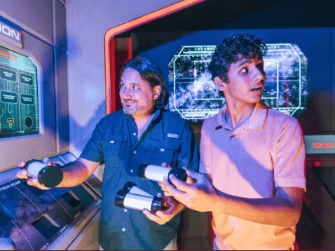 Two men play a sci‑fi arcade game, holding game controllers inside a neon-lit, high-tech arcade cabin with holographic screens behind them.