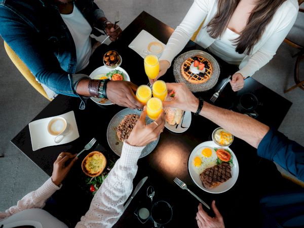 A group of friends cheers with orange juice at a table full of varied plates and desserts, celebrating together.