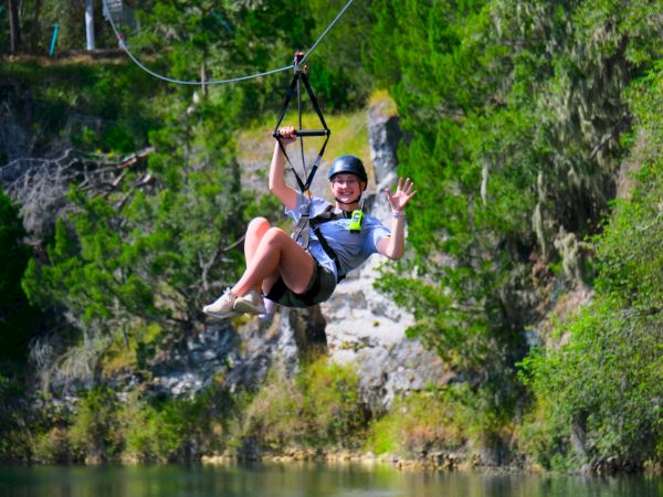 Person on a zip line over a lake, smiling and waving while wearing a helmet, cords and trees in the background.
