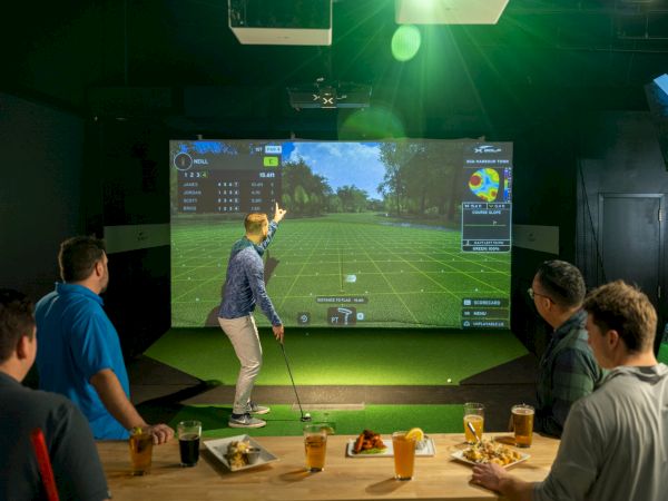 People are watching a large-screen golf game projections; a person is swinging while others sit with drinks and snacks in a dim room.
