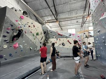 A busy indoor rock gym with climbers on angled walls and holds, belaying, chalk dust, and road-humped mats.