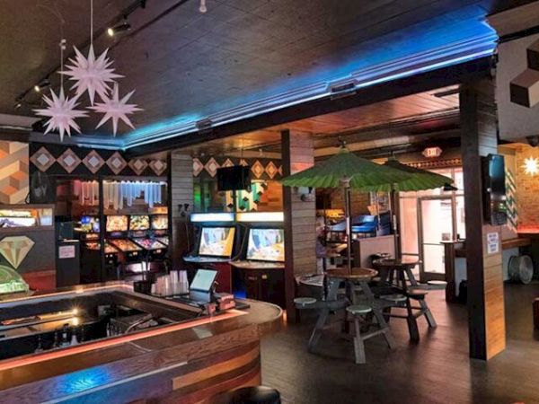 A retro arcade with pool tables, arcade machines, neon lighting, and quirky geometric decor, plus hanging paper stars and cozy seating, fun vibes.